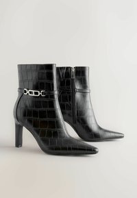 Bottines noires à bout pointu, texture en relief crocodile, fermeture zippée et accent en métal argenté sur la sangle de la cheville.