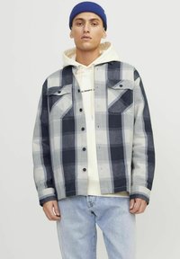 Camicia a quadri grigia, blu navy e bianca con due tasche sul petto, chiusura con bottoni e vestibilità comoda, indossata sopra un felpa color crema.