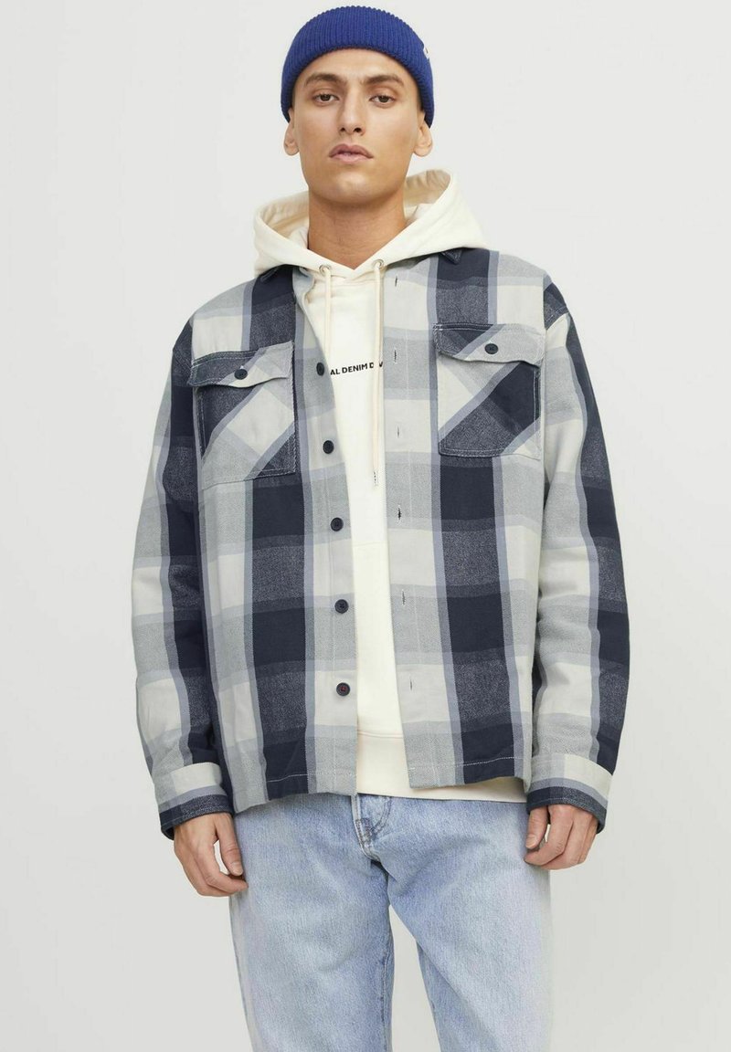 Camicia a quadri grigia, blu navy e bianca con due tasche sul petto, chiusura con bottoni e vestibilità comoda, indossata sopra un felpa color crema.