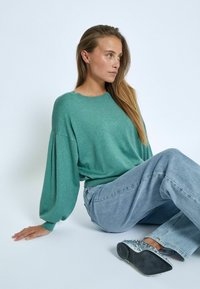 Green, textured sweater with dropped shoulder seams and subtle ribbing at the cuffs, paired with light blue jeans and embellished shoes.