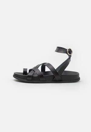 Sort flad sandal med tå-loop, flere remme over foden og ankelrem med sølvspænde, vist mod en simpel hvid baggrund.