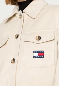 Kremowa kurtka z mieszanki wełny z kołnierzykiem, z dwiema przednimi kieszeniami, metalowymi guzikami oraz naszywką z logo "TOMMY JEANS" w kolorach niebieskim, czerwonym i białym.