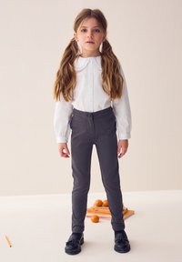 Biela košeľa s dlhým rukávom a volánovým golierom v kombinácii so sivými úzkymi nohavicami. Oblečenie dopĺňajú čierne topánky. Jednoduchý, čistý dizajn.