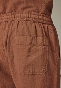 Pantaloni marroni con texture, dotati di una vita arricciata e dettagli di tasche laterali. Il tessuto presenta un discreto motivo a griglia e una vestibilità comoda.