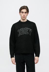 Pulover negru tricotat, cu un logo mare "OBEY" alb pe piept, guler rotund și mâneci largi. Purtat cu pantaloni închiși la culoare.