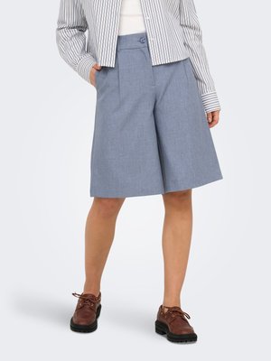 Kvinne iført knelang lyseblå skreddersydde shorts, stripete blazer, hvit topp og brune snøresko, står med en hånd i lommen.
