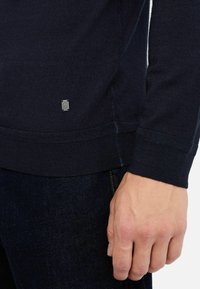 Maglione navy con polsini e orlo a coste, realizzato in morbido materiale a maglia. Presenta un piccolo tag in metallo sul lato e una texture liscia.