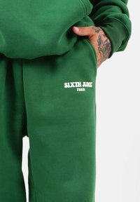 Groene joggerbroeken van zachte stof, met op de linkse zak een witte logo-tekst "SIXTH JUNE PARIS". Glad textuur en een relaxte pasvorm.