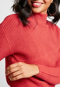Pull en maille côtelée rouge avec un col montant et un motif texturé. Le tissu semble doux, et les manches sont ajustées.