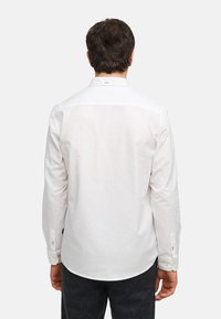 Chemise blanche à manches longues avec poignets à boutons, texture lisse, ourlet arrondi et un détail de col subtil. Portée avec un pantalon foncé à motifs.