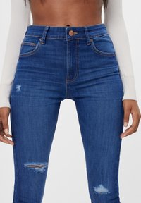 Jeans de tiro alto en azul, fabricados en denim, con un diseño ajustado, áreas ligeramente desgastadas, costuras naranjas y bolsillos frontales clásicos.