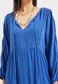 Modrá dlouhá šatová šaty s výstřihem do V a strukturovanou látkou. Obsahují krajkové detaily a uvolněný střih. Doplněné o vrstvený zlatý náhrdelník.