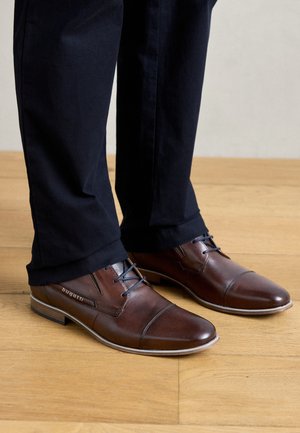 Chaussures de costume en cuir marron avec un design à bout fleuri, lacets bleus et semelle beige, assorties à un pantalon sombre, sur un sol en bois.