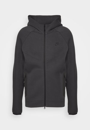 Mörkgrå zip-up hoodie med hög krage, raglanärmar, sidofickor och subtilt präglat logotyp på bröstet. Mjuk, slät tygtextur.