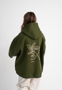 Hoodie vert olive au design surdimensionné. Présente un dessin linéaire minimaliste d'un visage et de feuilles au dos. Tissu doux et texturé.