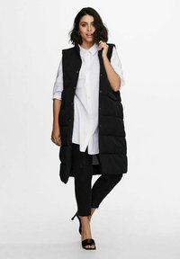 Juodas kilpinis ilgas liemenė su aukštu apykakle, dėvima ant baltos marškinėlių su sagomis ir juodų liesų džinsų. Turi spaudinius ir šonines kišenes.