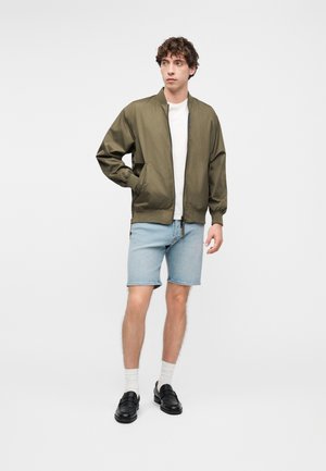 Mladi muškarac obučen u maslinasto zelenu jaknu na patentni zatvarač, bijelu majicu, svijetloplave traper kratke hlače, bijele čarape i crne mokasinke, stoji s jednom rukom u džepu.