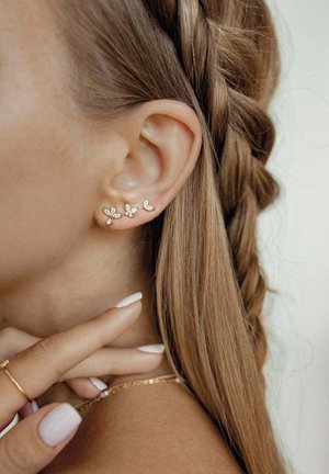 Gold-colored ear cuffs with leaf-like designs, featuring small crystals. Hair braided on one side, finger touching the neck.
