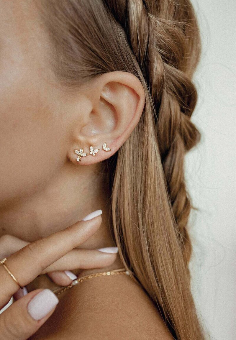 Pendientes de oreja color dorado con diseños similares a hojas, que cuentan con pequeños cristales. Pelo trenzado a un lado, dedo tocando el cuello.