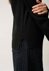 Schwarzer Strickpullover mit einem V-Ausschnitt am Saum, getragen über einem strukturierten, dunklen Faltenrock. Verfügt über Rippdetails an Ärmeln und Körper.