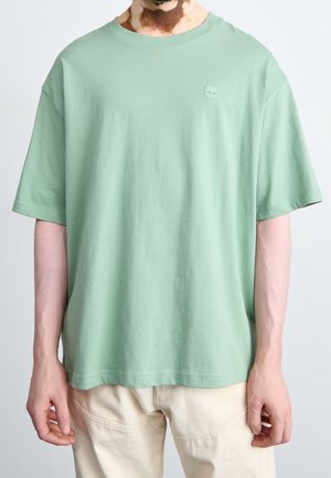 Person iført en lysegrøn kortærmet T-shirt med et lille hvidt trælogo på venstre bryst og cremefarvede bukser mod en ensfarvet baggrund.