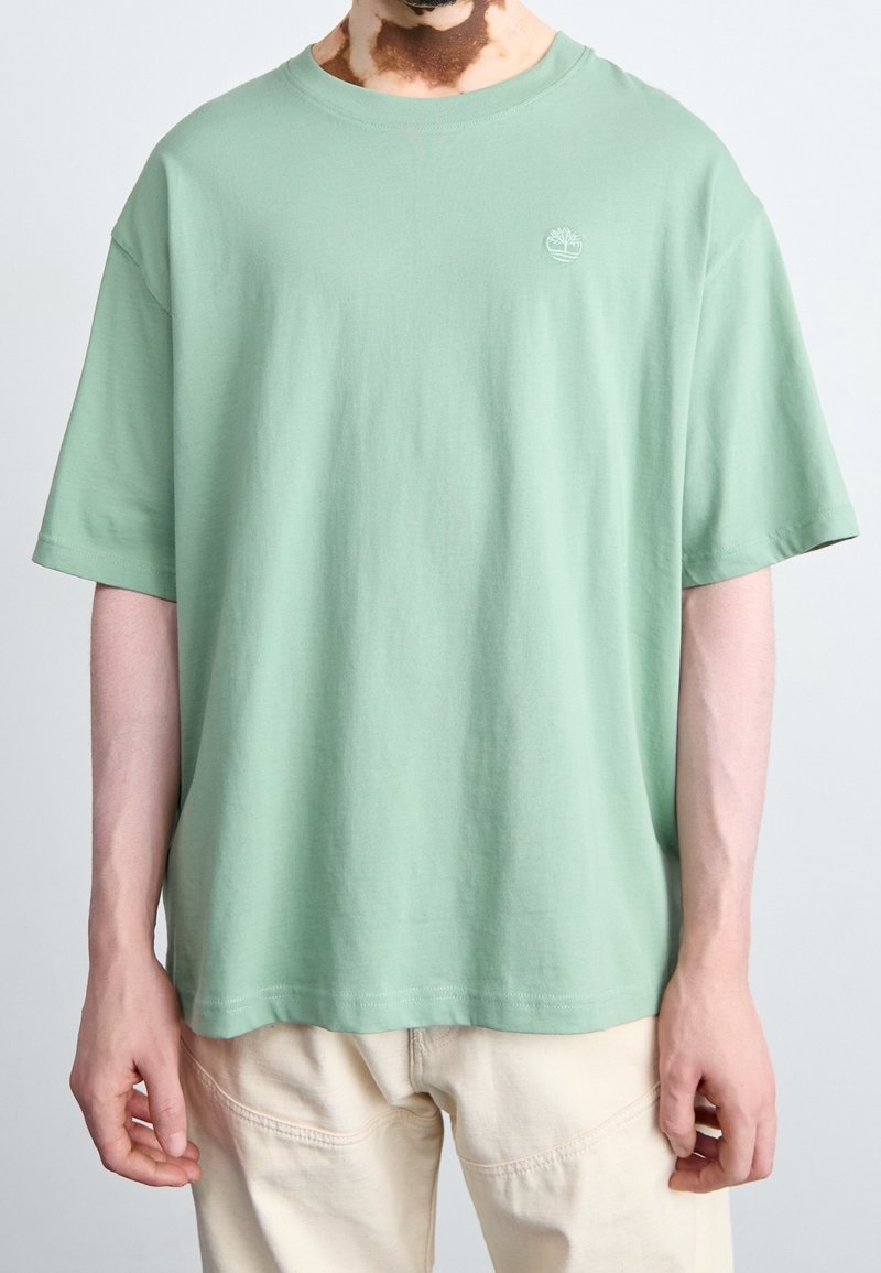 Personne portant un t-shirt vert clair à manches courtes avec un petit logo arbre blanc sur la poitrine gauche et un pantalon crème sur un fond uni.