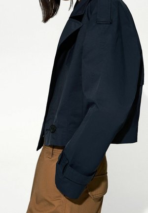 Veste courte bleu marine à la coupe structurée, avec grands revers et détails de boutons ; portée avec un pantalon beige. Texture lisse, silhouette moderne.