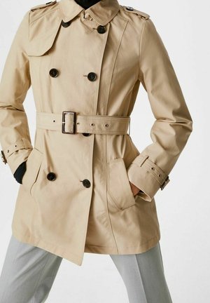 Beige dobbeltbrystet trenchcoat med bælte, sorte knapper og sidelommer båret over lysegrå bukser.