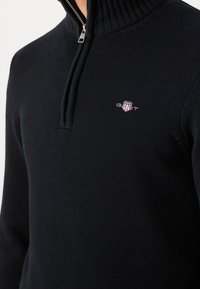 Pull-over noir en maille avec une fermeture éclair quart de zip et un petit logo GANT brodé sur la poitrine gauche.