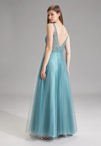 Abito blu aqua con gonna in tulle, corpetto con perline e design con profondo scollo a V sul retro. Presenta spalline sottili e una silhouette fluida. Tacchi argentati visibili.