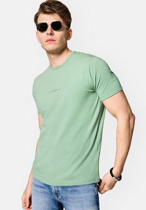 Jasnozielony t-shirt z krótkim rękawem wykonany z miękkiego materiału, z okrągłym dekoltem i subtelnym nadrukiem logo z przodu. Nosi się go z niebieskimi dżinsami.