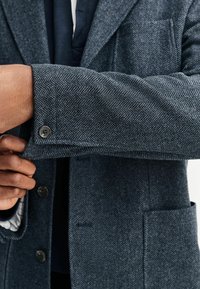 Dunkelblauer Blazer mit Fischgratmuster und strukturiertem Stoff; verfügt über knöpfbare Manschetten und eine Brusttasche, die auf maßgeschneiderte Designelemente hinweist.