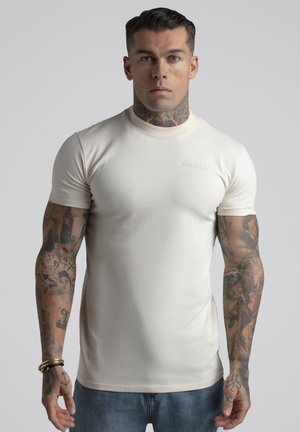 Mann mit Tätowierungen am Hals und an den Armen, der ein eng anliegendes, einfarbiges, kremfarbenes T-Shirt und blaue Jeans trägt und vor einem einfachen Hintergrund steht.