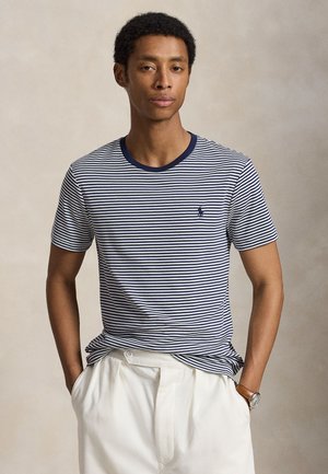 Homme portant un t-shirt rayé bleu marine et blanc avec un petit logo, un pantalon blanc et une montre-bracelet, debout les mains dans les poches.