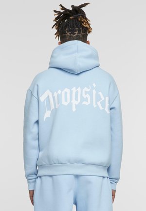 Persoană cu dreadlocks legate poartă un hanorac și pantaloni bleu, cu spatele spre privitor, având text gotic alb "Dropsize" pe hanorac.