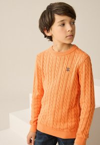 Oranger Kabelstrickpullover mit gerippten Bündchen und Saum, ausgestattet mit einem kleinen navyblauen Logo auf der Brust, kombiniert mit dunkelblauen Jeans.