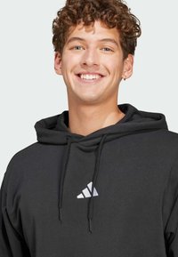 Crna trenirka s kenguruvim džepom, s bijelim Adidas logom na prsima, kapuljača s vezicom i mekani tekstil.