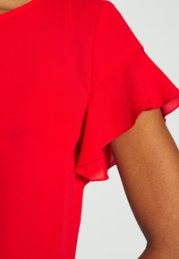 Top in tessuto leggero rosso con design a maniche a goccia fluttuanti. Texture liscia, scollatura rotonda, con dettagli di cucitura sulla spalla.