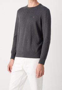 Homme portant un pull à col rond manches longues gris foncé avec un petit logo rouge, blanc et bleu sur la poitrine et un pantalon blanc.