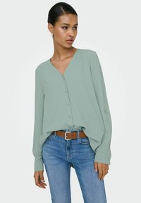 Femme aux cheveux plaqu és en arrière portant des boucles d'oreilles argentées en forme d'anneau, un chemisier vert clair boutonné, une ceinture marron et un jean bleu, regardant sur le côté.