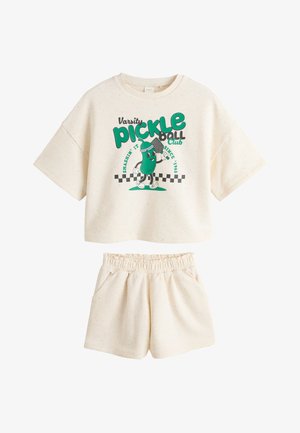 Młode kręcone dziecko w kremowej koszulce i szortach do pickleball stojące przy rustykalnych białych drzwiach z kwiatami w pobliżu.