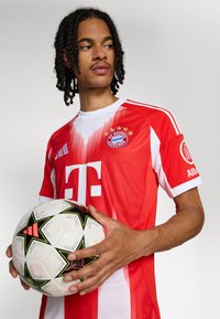 Maillot rouge et blanc avec le logo du FC Bayern Munich et les détails de parrainage ; le joueur tient un ballon de football blanc avec des accents noirs et verts.