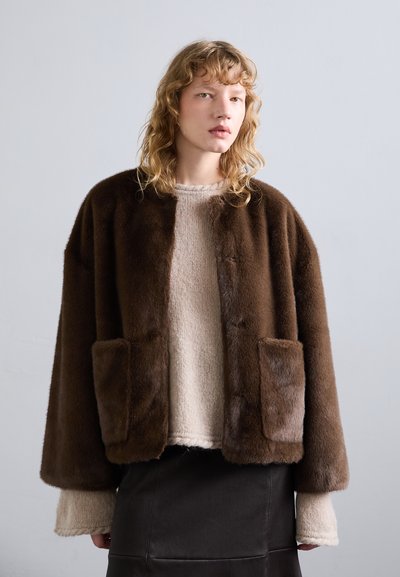 Brun fuskpälsjacka med en avslappnad passform, två framfickor och en mjuk textur, bärs över en ljusbeige tröja och en svart kjol.