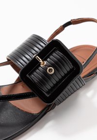 Sandalo nero caratterizzato da una cintura lucida con una grande fibbia rettangolare, interno morbido color marrone e dettagli di design minimalisti.