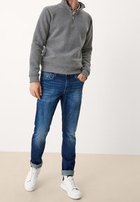 Felpa grigia con zip e finitura texture, abbinata a jeans blu in denim con un effetto sbiadito. Sneakers bianche completano l'outfit.