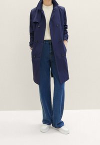 Tmavě modrý trenčkot s dvojitým zapínáním na knoflíky, velkými knoflíky a bočními kapsami. Nošený přes krémový pletený top a modré džíny s širokými nohavicemi.