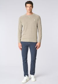 Maglione beige chiaro con scollatura rotonda e polsini a coste, abbinato a pantaloni blu scuro e sneakers bianche. Design semplice con un accento del logo.