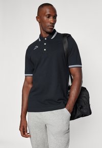 Polo nero con colletto bianco e strisce sulle maniche, dotato di tre bottoni e un piccolo logo sul petto, abbinato a pantaloni felpati grigi.