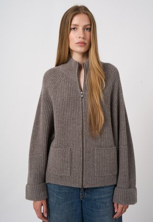 Frau mit langen blonden Haaren, die einen grauen, gerippten Strick-Zipperpullover mit großen Bündchen und Fronttaschen trägt, kombiniert mit blauen Jeans, vor weißem Hintergrund stehend.