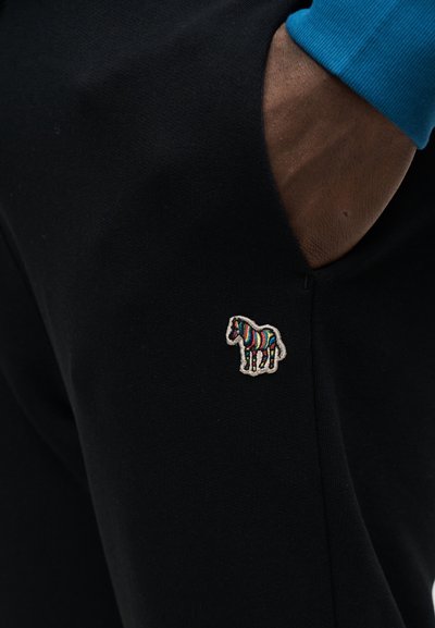 Χέρι στην τσέπη μαύρου παντελονιού που δείχνει πολύχρωμο κεντητό μοτίβο ζέβρας σε μαύρο ύφασμα, άτομο που φορά μπλε μακρυμάνικο μπλουζάκι.